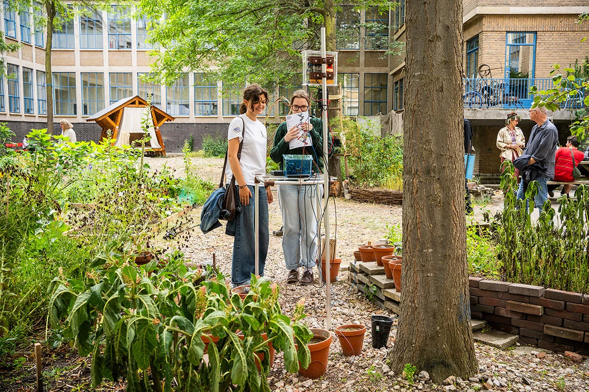 Installation, comprised by metal tubing, electronics and vases with mud all wired together, seen from behind as two visitors interact with it. 
