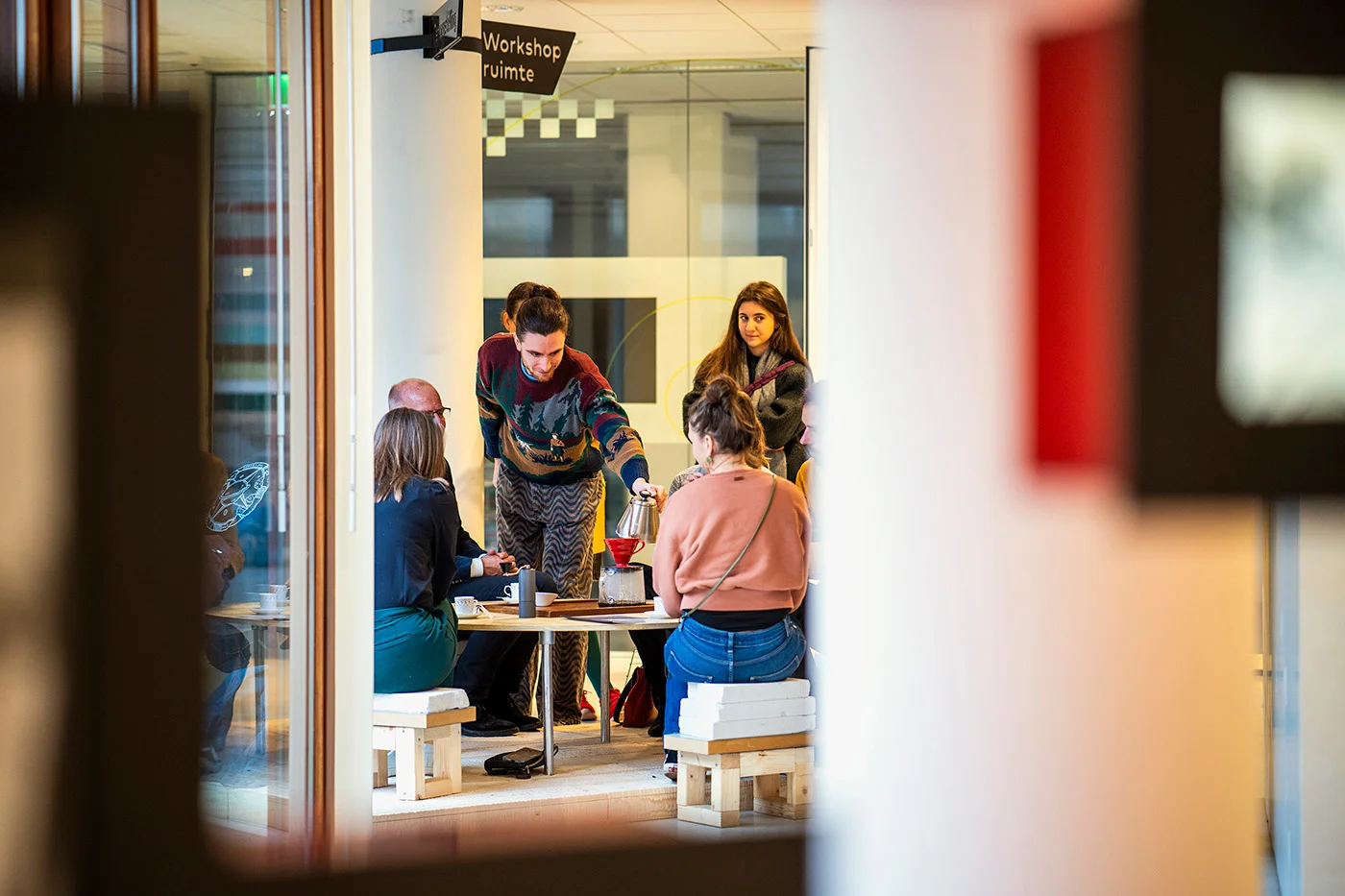 View of the same group as before, sitting around the table, partially hidden behind a pilar, from further away. 