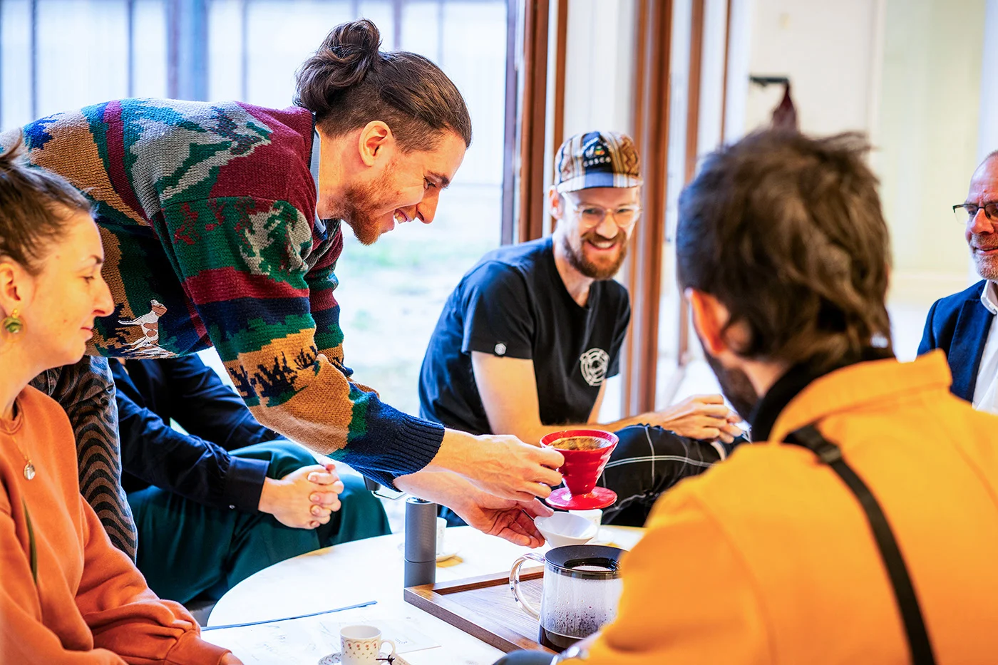 Medium shot of a group of 5 people sat around a low round table, smiling as coffee is prepared.