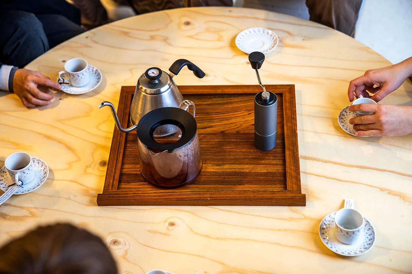 Top view of a round wooden table. One can see people's hands holding cups with coffee and a tray with a kettle, a garrafe, half filled with coffee, and a grinder.